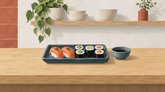 Illustration til sushirestauranter i Danmark: nigiri og maki på en blå bakke med en lille skål sojasauce på en lys trædisk.
